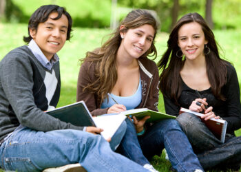 A group of three young people, two women and one man, sit on grass outside with notebooks and pens, smiling at the camera. They appear to be students studying together in a park or campus setting with trees and greenery in the background, possibly discussing plans for paying off student loans. | TraceLoans