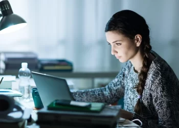 A woman with a long braid works intensely on her laptop at a cluttered desk with books, a water bottle, and other items, searching for information on how to find student loans. A desk lamp illuminates the scene in an otherwise dimly lit room. | TraceLoans