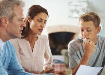 A middle-aged man and woman sit with a teenage boy, all appearing to focus on a document the boy is holding. The boy looks thoughtful, his chin resting on his hand, while the woman speaks about student loan refinance options and the man listens attentively. They are in a cozy, well-lit living room. | TraceLoans