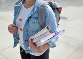A person wearing a light denim jacket and a white shirt with small orange shapes holds multiple books in one hand and has a gray backpack on their shoulders. They are walking on a sidewalk with earphones in, possibly contemplating the weight of student loans. | TraceLoans