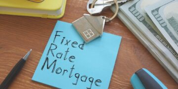 A desk with a blue sticky note labeled "Fixed-Rate Mortgage." Nearby are house keys on a keychain shaped like a house, a stack of hundred-dollar bills, a pen, a stapler, and a few notebooks. | TraceLoans