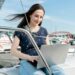 A woman with long dark hair, wearing a blue patterned shirt and blue jeans, sits on a sailboat and works on her laptop. The backdrop features a marina with several sailboats docked under a clear blue sky. She’s probably researching the best bank for digital nomads to manage her finances efficiently. | TraceLoans