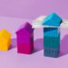 Three model houses placed in ascending order of size on a purple background. The smallest house is yellow, the medium house is magenta, and the largest house—a stack of three blue houses with U.S. dollar bills on its roof—symbolizes fixed rate stability in housing investments. | TraceLoans