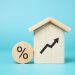 A wooden house with an upward arrow symbol stands next to a wooden disc featuring a percentage sign, set against a light blue background, representing increasing average mortgage rates or property value. | TraceLoans