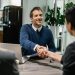 A man in a blue sweater is smiling and shaking hands with another person across a table, likely discussing business loans. A woman sits next to them, while an open laptop captures their focus. In the background, plants and a refrigerator enhance the professional office setting. | TraceLoans