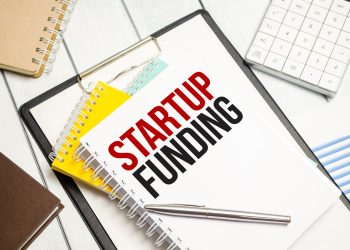 A desk with a notebook labeled "STARTUP FUNDING" in bold black and red letters is neatly arranged with a pen, a calculator, and other notebooks. A nearby paper titled "Startup Loans" adds an extra layer of planning on the white wooden surface. | TraceLoans
