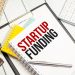 A desk with a notebook labeled "STARTUP FUNDING" in bold black and red letters is neatly arranged with a pen, a calculator, and other notebooks. A nearby paper titled "Startup Loans" adds an extra layer of planning on the white wooden surface. | TraceLoans