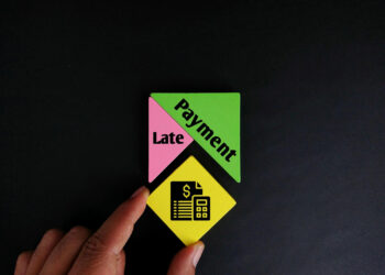 A hand holds a yellow square with icons of a dollar sign, receipt, and calculator below two joined triangles labeled "Late Payment" on a black background. | TraceLoans