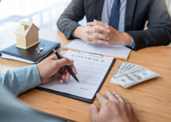 Two people sit at a table; one signs a contract while the other has folded hands. A small house model, clipboard, and stack of hundred-dollar bills are on the table, suggesting a real estate or financial agreement. | TraceLoans